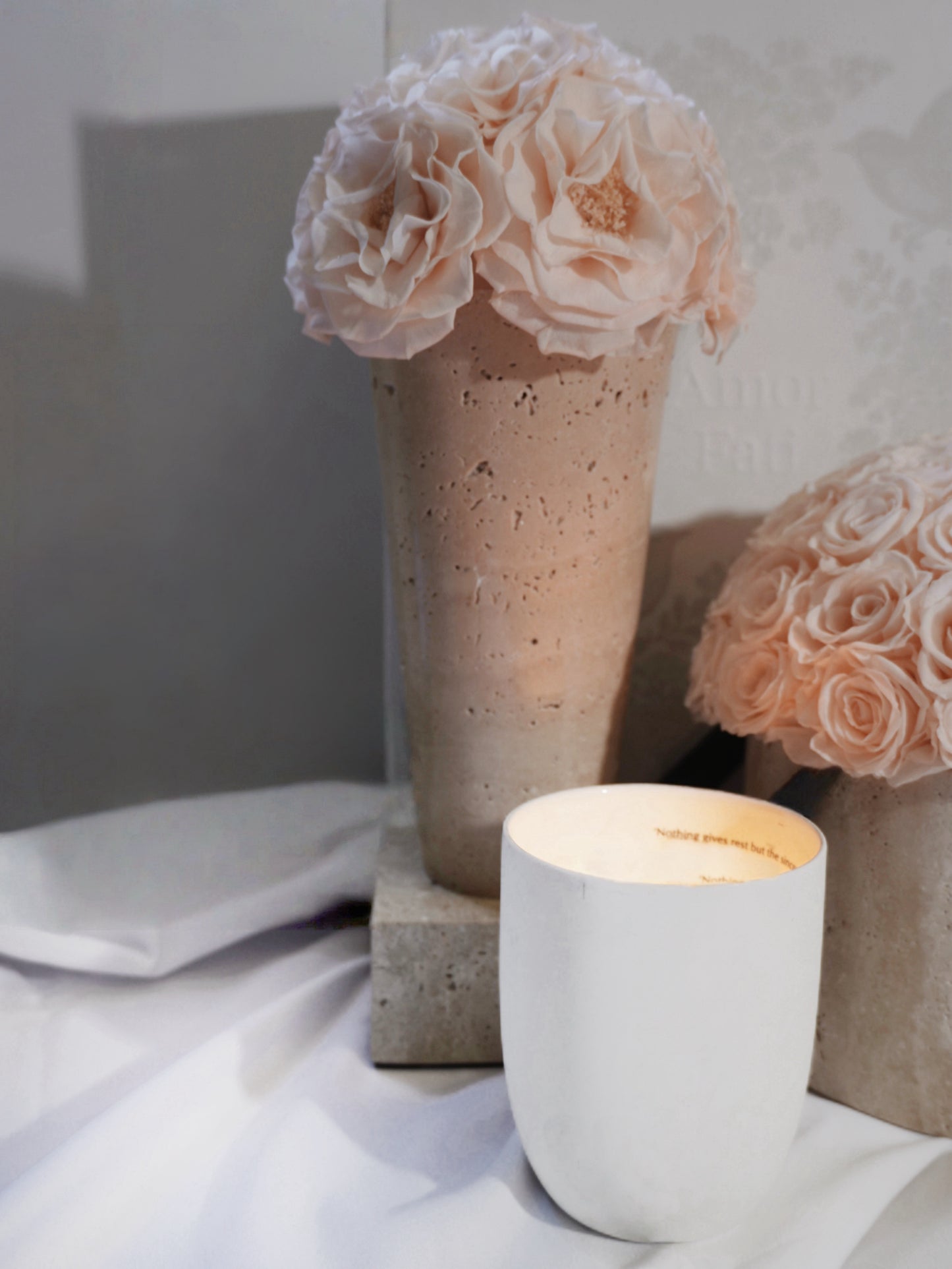 Two geometric travertine vases with light pink preserved roses are placed on a soft white cloth. Beside them is a white candle, emitting a warm and elegant atmosphere, which is a great choice for home decoration.