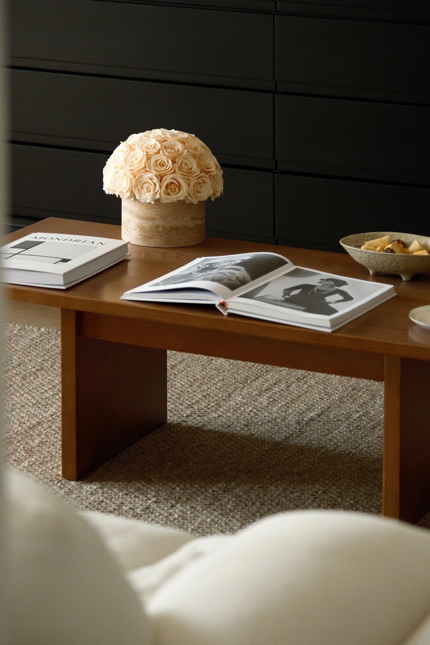 A bunch of beige eternal roses in a travertine container sits on a wooden coffee table. Around it are open magazines and a bowl of snacks, creating a warm and inviting living room scene.