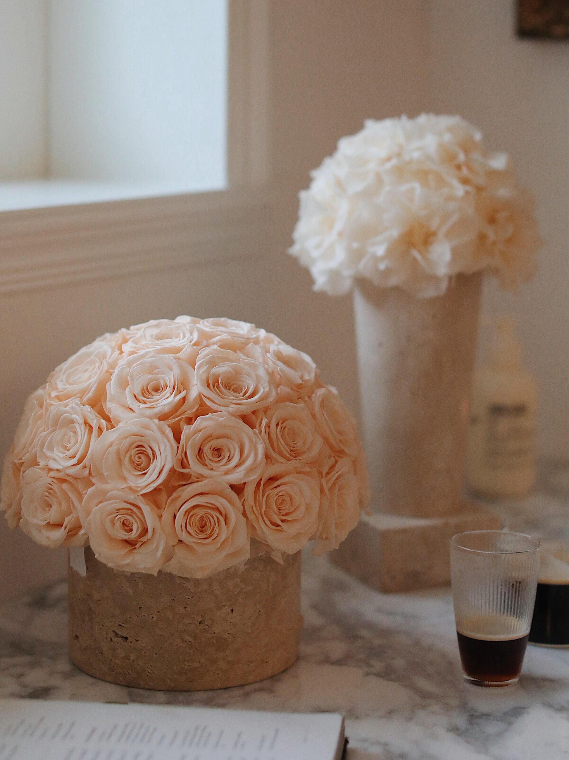 On a marble surface, a large bouquet of  preserved roses in a travertine container is accompanied by a glass of iced coffee, exuding a sense of elegance and comfort.