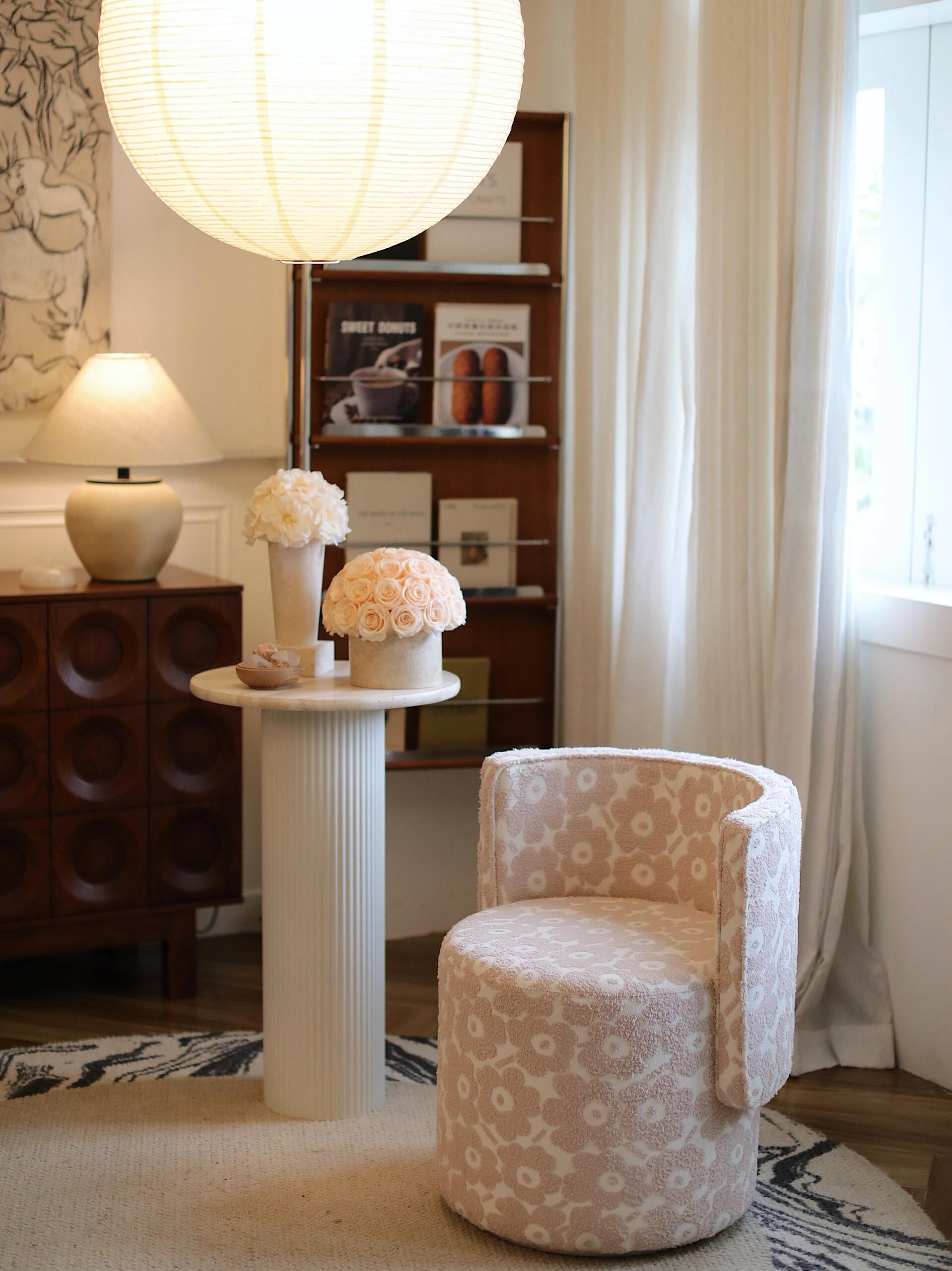 In a softly lit room, a patterned chair sits beside a white columnar side table. On the table are two small arrangements of preserved roses, accompanied by a small decorative item.