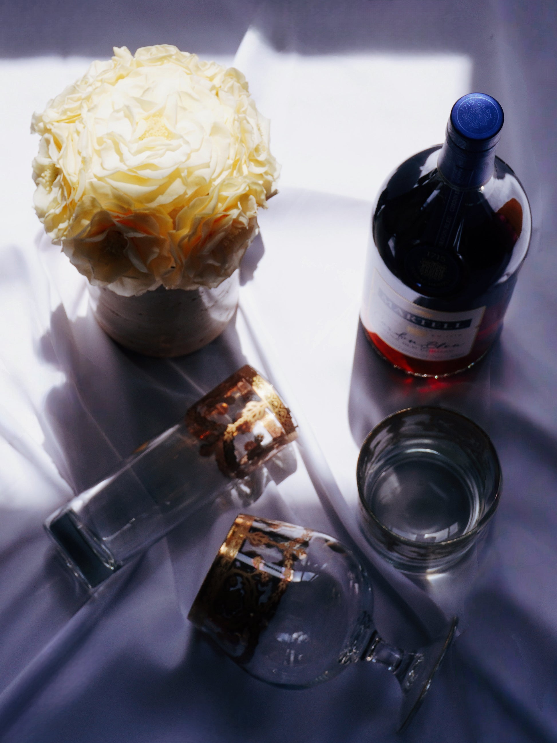 A cluster of preserved flowers in a travertine vase is positioned beside a wine bottle and elegant wine glasses on a soft white cloth, bathed in warm light, exuding a cozy and stylish home ambiance.