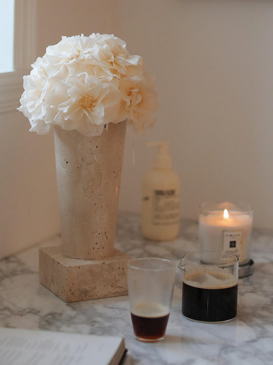 A geometric - shaped travertine vase holds a bouquet of white preserved roses. Alongside are a lit candle and two glasses of beverages, with a book beneath. This setup creates a serene and elegant atmosphere, perfect for enhancing home decor.
