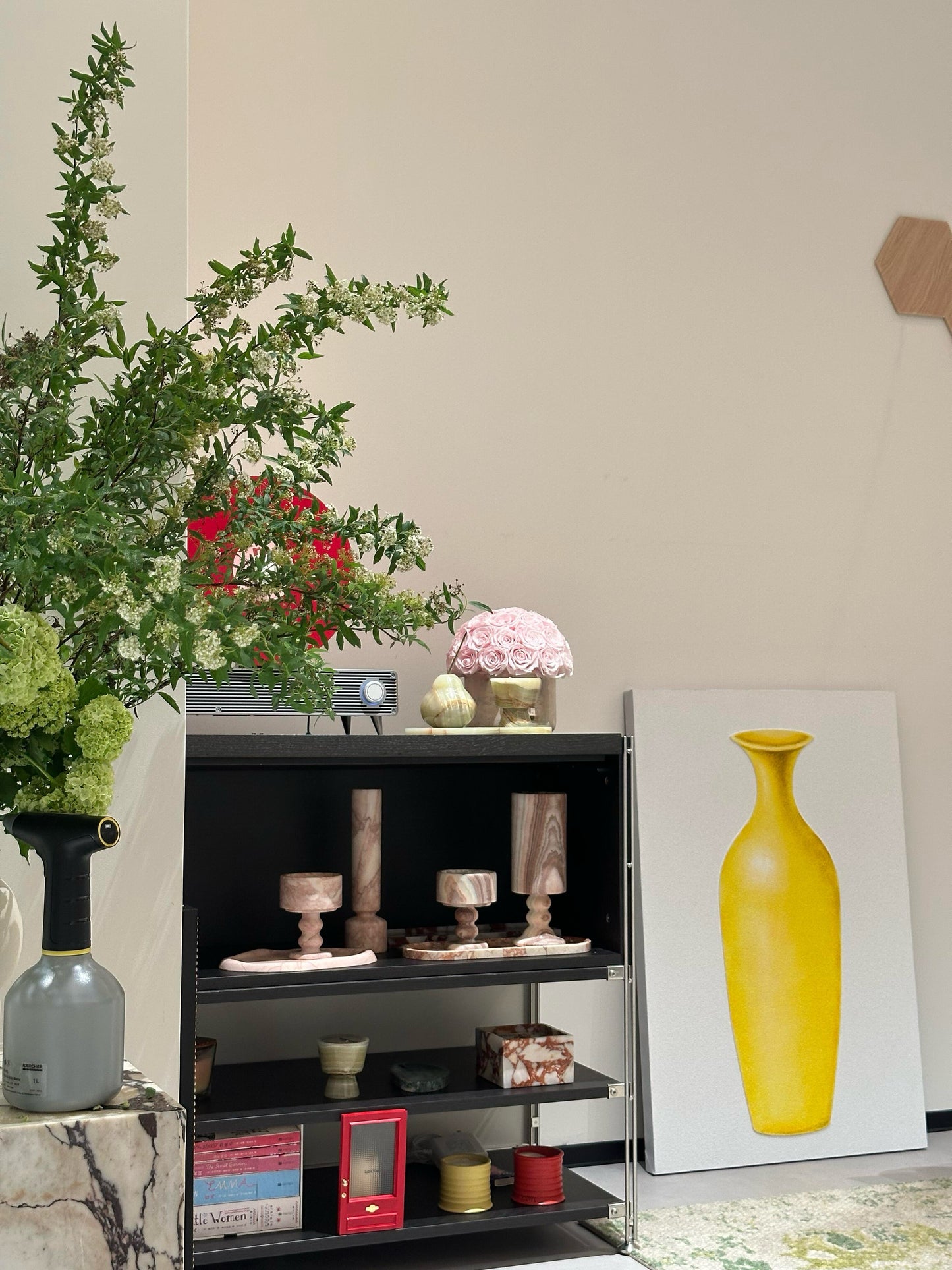 The pink preserved roses in a yellow travertine container stand out on the shelf. They blend with other decorative items, adding a soft and elegant touch, and become a highlight in the room's decor.