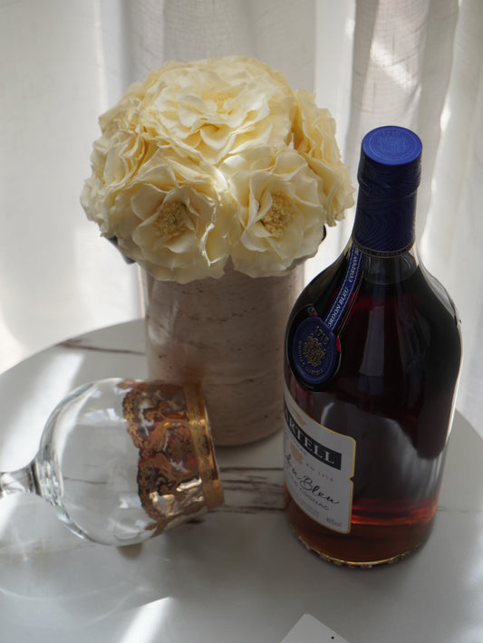 A bouquet of elegant white eternal flowers in a natural travertine vase stands gracefully. Paired with a bottle of liquor and a stylish glass, it creates a sophisticated and timeless scene, perfect for adding a touch of luxury to any space.