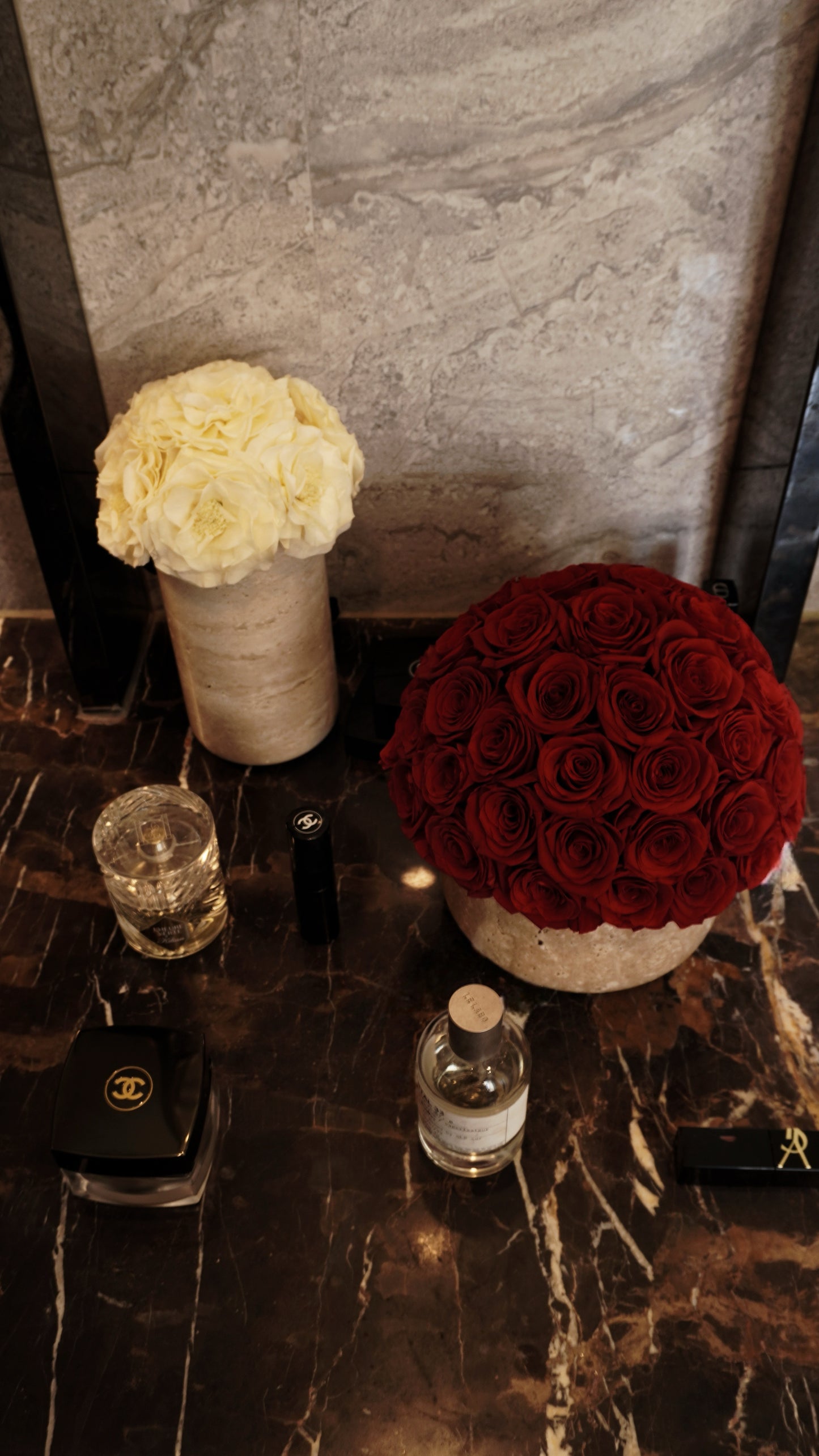 On the luxurious marble surface, a bouquet of deep red preserved roses and a cluster of white preserved flowers in travertine containers are placed alongside elegant perfume bottles. They bring a blend of romance and refinement, becoming the eye - catching decor highlights of the space.