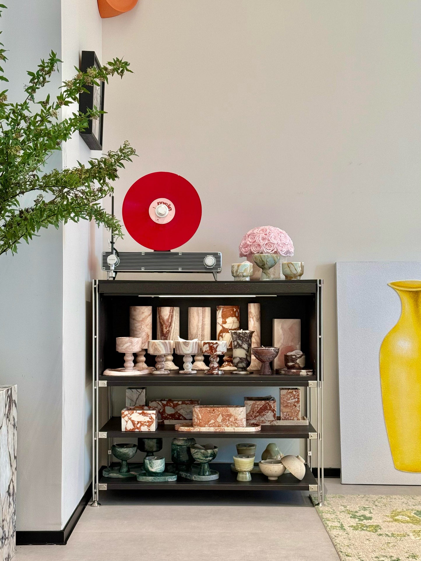 On the shelf, a bunch of pink preserved roses in a yellow travertine container is placed among various artworks and decorations. It acts as a delicate and soft - toned embellishment, breaking the monotony of the space and adding a touch of natural and romantic charm.
