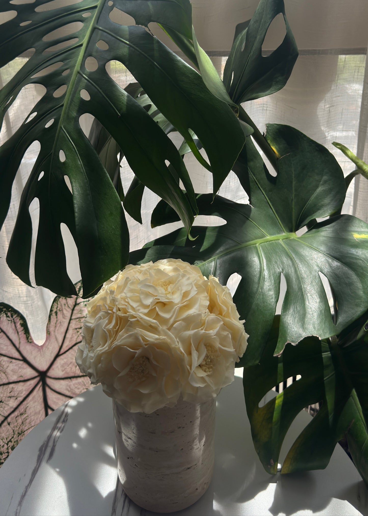 A bunch of preserve  rose in a natural travertine vase is set against a backdrop of lush monstera leaves. The contrast between the delicate flowers and the bold greenery creates a visually appealing scene, bringing a touch of nature's beauty and tranquility indoors.