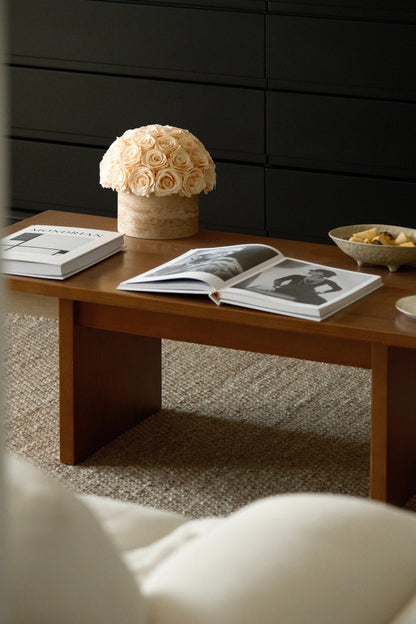 A bunch of beige eternal roses in a travertine container sits on a wooden coffee table. Around it are open magazines and a bowl of snacks, creating a warm and inviting living room scene.