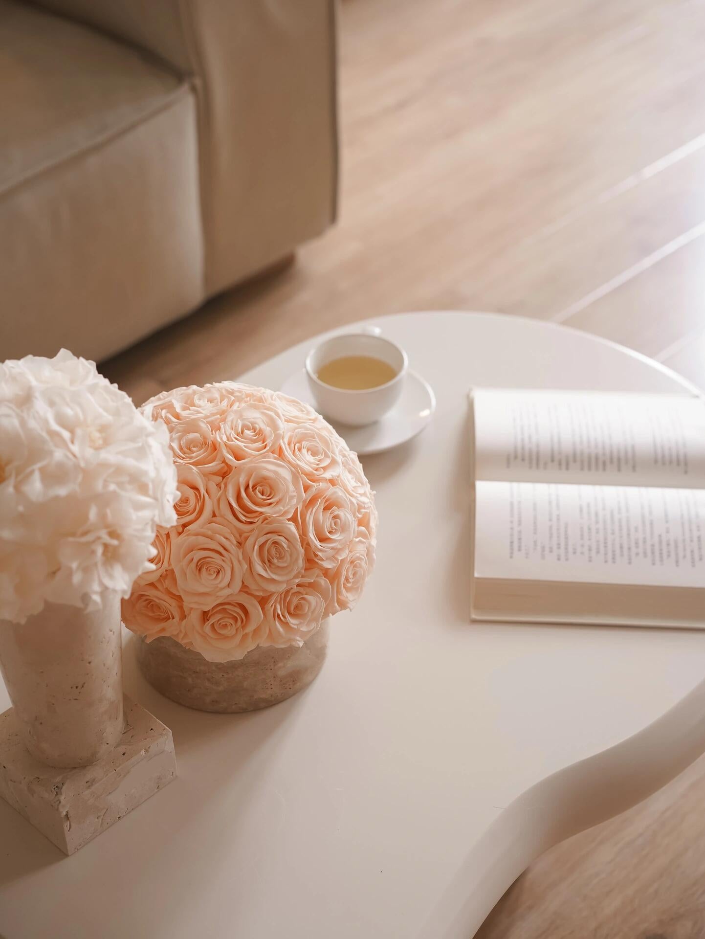 On a white coffee table, there are two pots of preserved roses in different shades, a cup of tea and an open book, creating a leisurely and elegant home atmosphere.