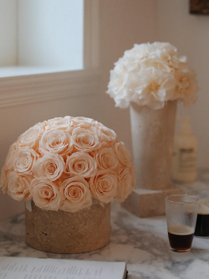 On a marble surface, a large bouquet of  preserved roses in a travertine container stands out. Nearby is a glass of iced coffee, bringing a touch of elegance and refreshment to the scene.
