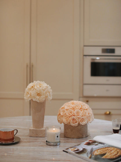 On a marble dining table, there are two pots of preserved roses in different styles, accompanied by a cup of coffee, a lit candle, and a plate of pastries, creating a warm and elegant atmosphere