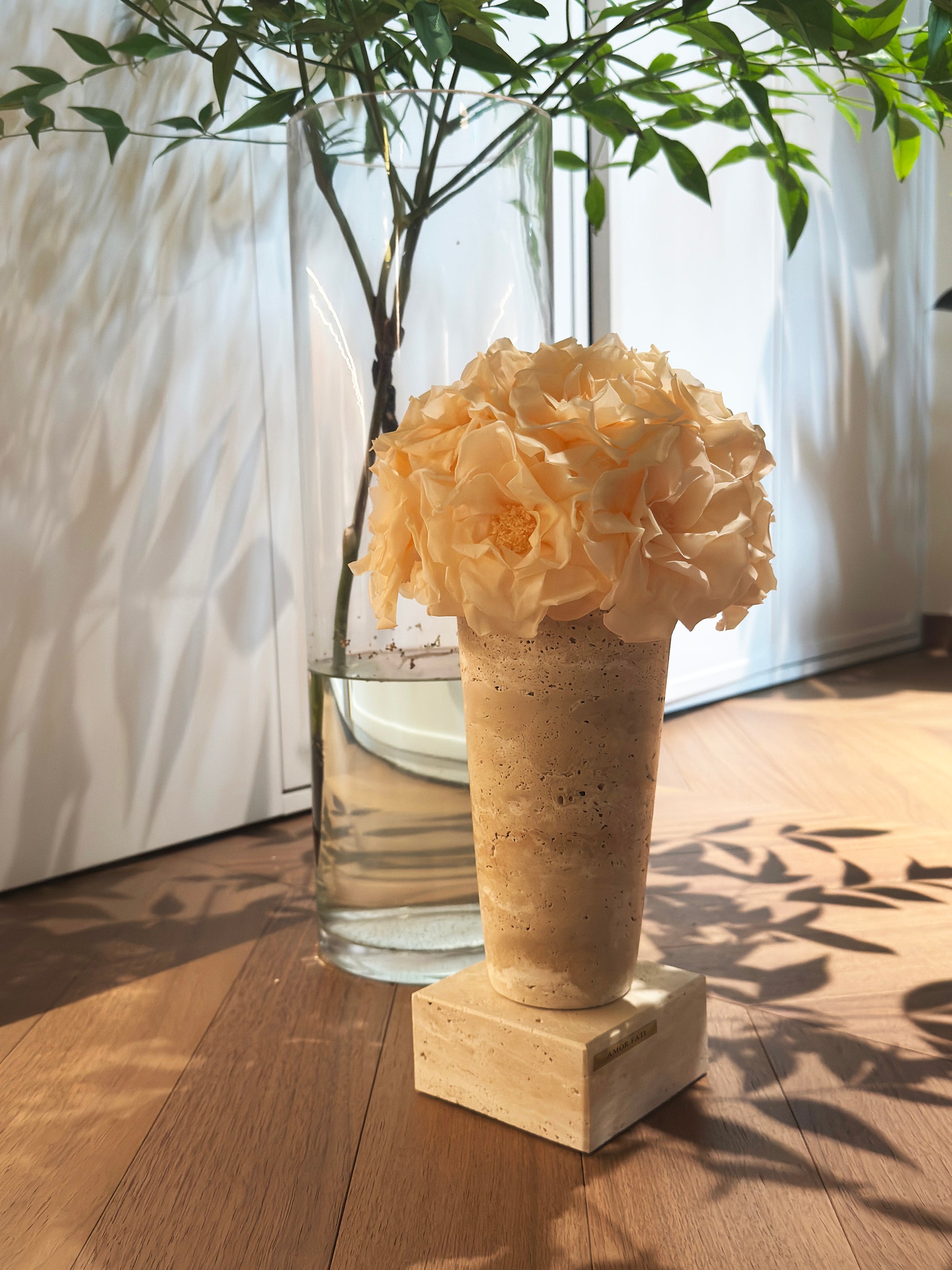 A geometric - shaped travertine vase with a cluster of light - colored preserved roses sits on a wooden floor. Behind it is a glass vase with green plants, allowing natural light to cast shadows, creating a warm and stylish decor scene.