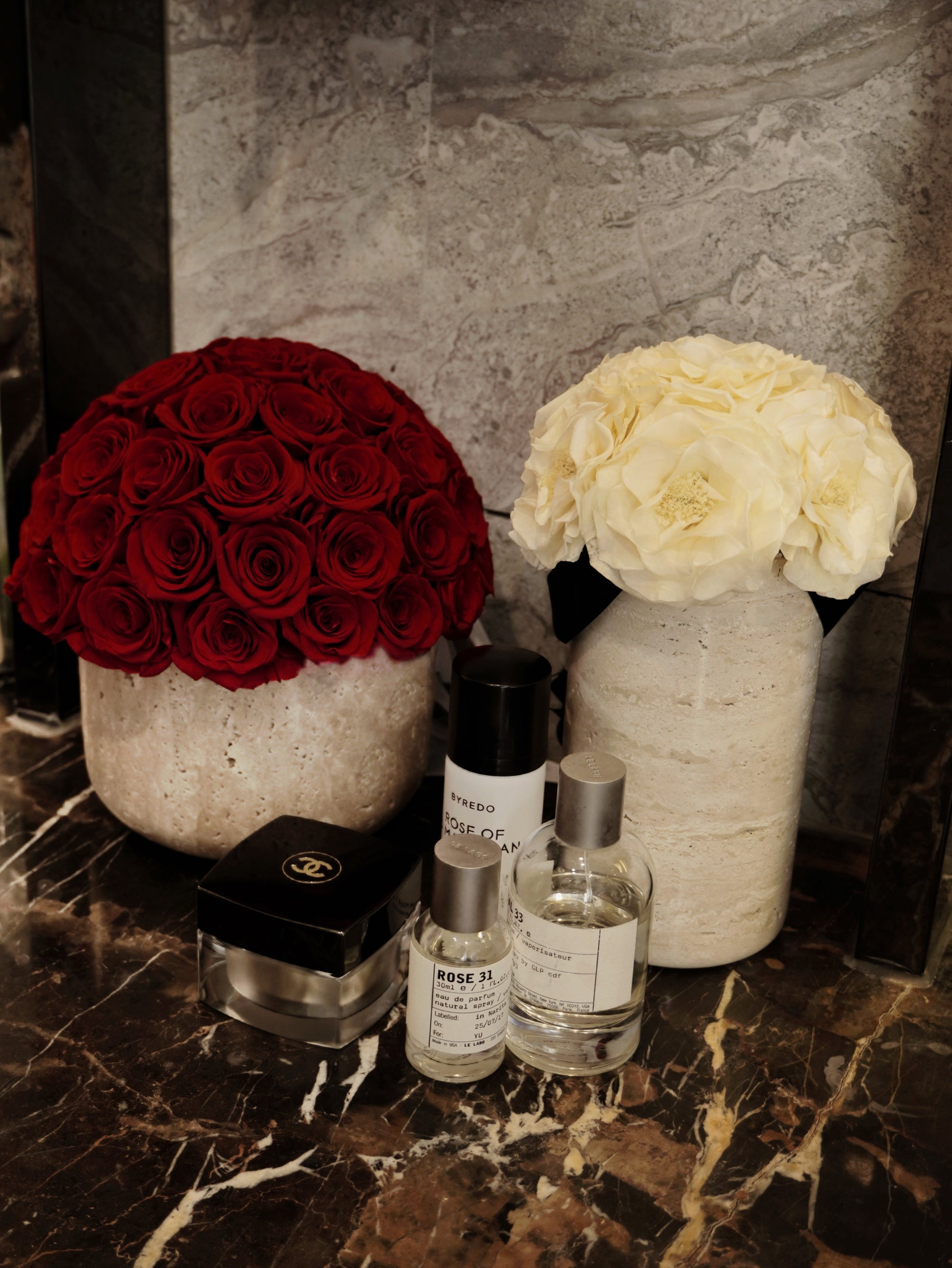 On the luxurious marble surface, a bouquet of deep red preserved roses and a cluster of white preserved flowers in travertine containers are placed alongside elegant perfume bottles. They bring a blend of romance and refinement, becoming the eye - catching decor highlights of the space.