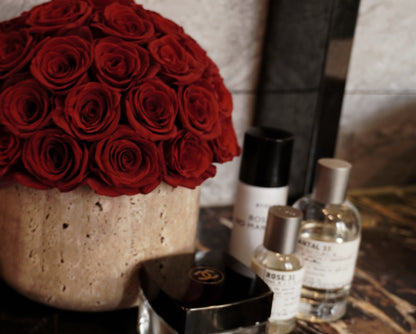 A gorgeous cluster of deep red preserved roses in a travertine container stands out beside elegant perfume bottles. This combination not only adds a touch of romance and luxury to the space but also creates a visually appealing focal point for any room.