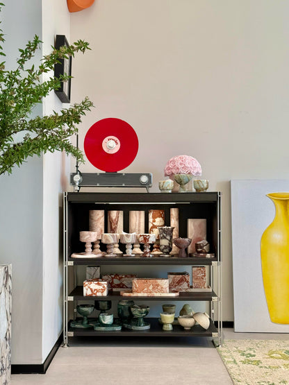 On the shelf, a bunch of pink preserved roses in a yellow travertine container is placed among various artworks and decorations. It acts as a delicate and soft - toned embellishment, breaking the monotony of the space and adding a touch of natural and romantic charm.