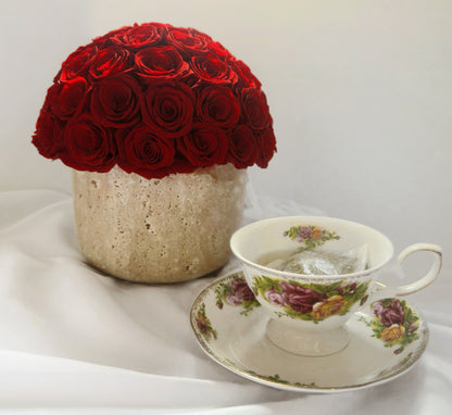 A striking arrangement of red preserved roses in a natural travertine container sits elegantly beside a vintage - styled teacup with floral patterns. This combination creates a warm and refined atmosphere, making it a charming decorative piece for any setting.