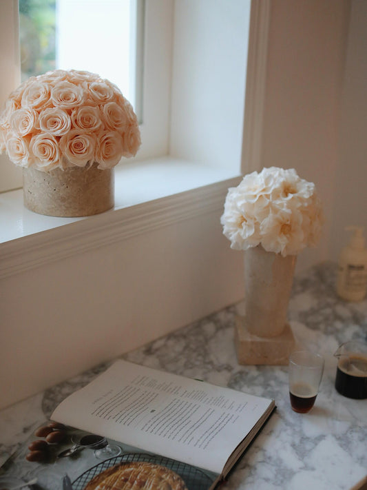 On the marble windowsill, two bouquets of beige eternal roses in travertine containers are placed. Next to them is an open book, a glass of drink, creating a cozy and elegant home corner.
