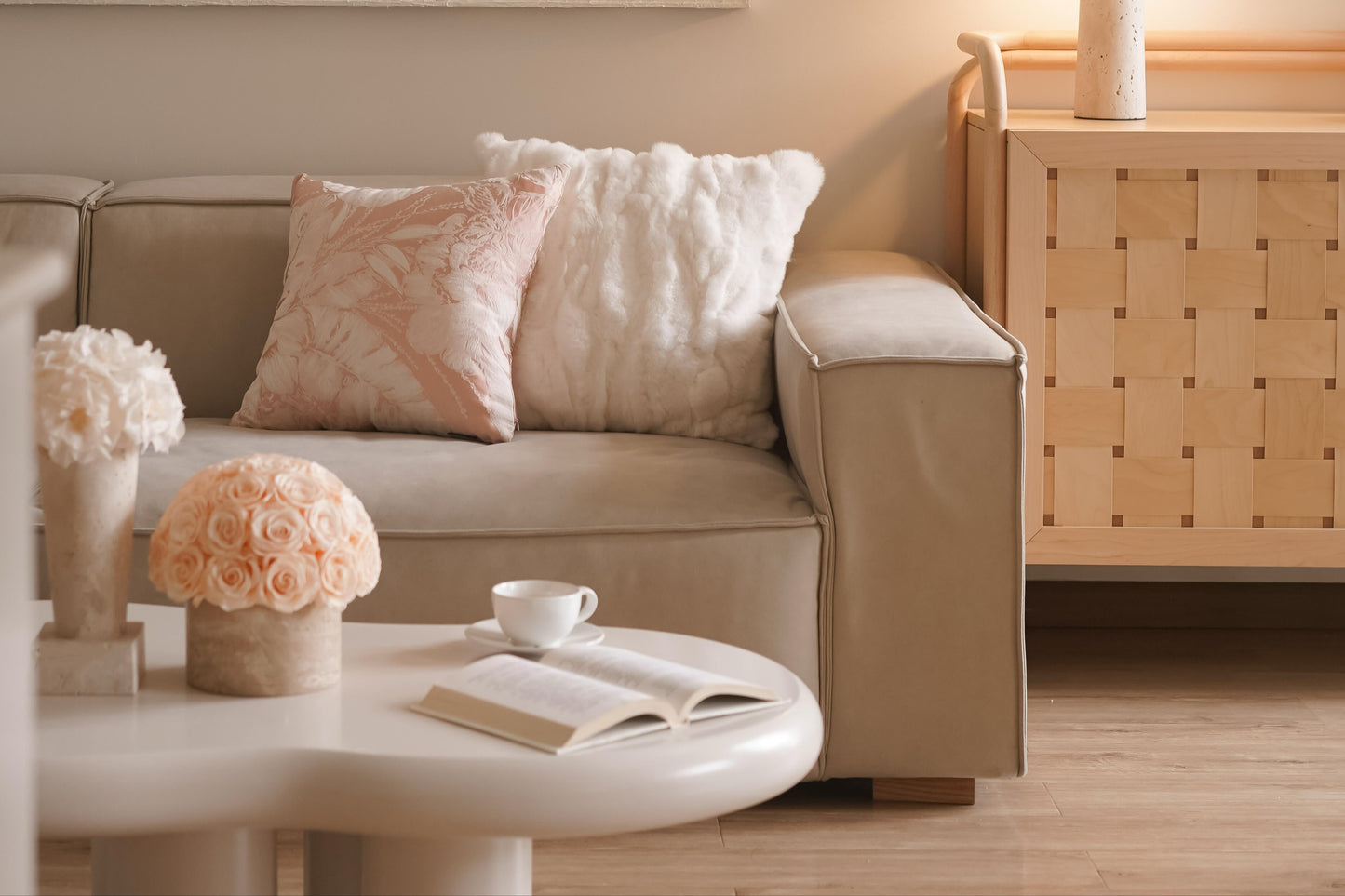 A beige sofa adorned with soft cushions. On the round coffee table in front, there are two pots of preserved roses, a cup of coffee and an open book, crafting a comfortable and inviting living space.