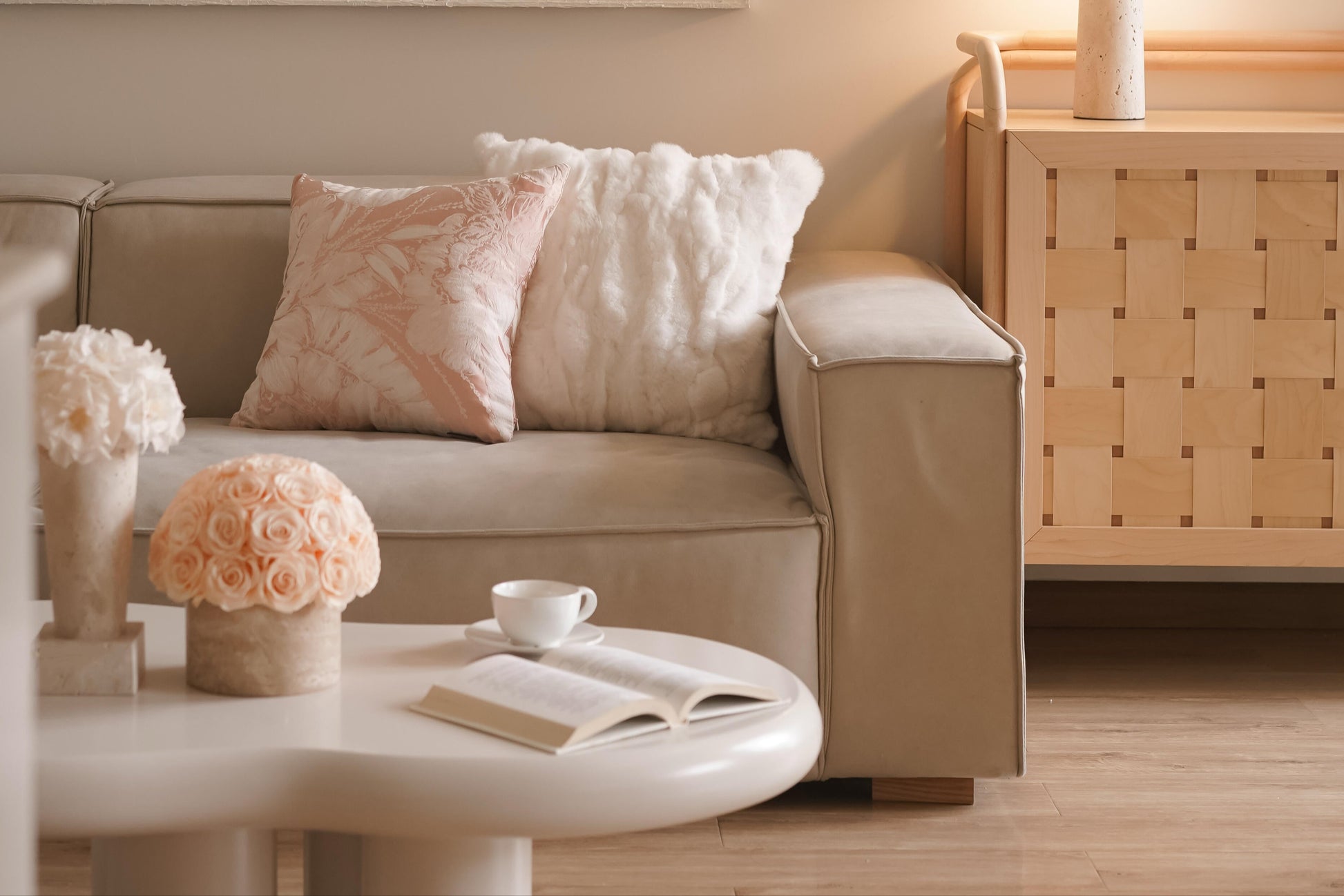 A beige sofa adorned with soft cushions. On the round coffee table in front, there are two pots of preserved roses, a cup of coffee and an open book, crafting a comfortable and inviting living space.