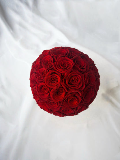 A sphere composed of vibrant red preserved roses rests on a white fabric background, exuding elegance and charm.